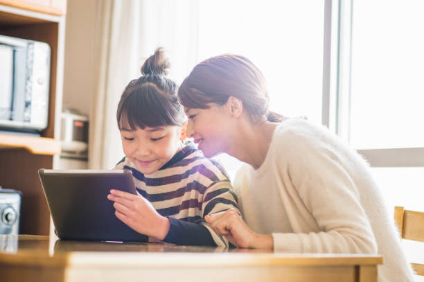 A woman and a child sit at a table, looking at a tablet together, with sunlight streaming through the window.