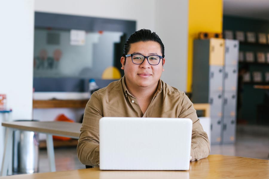 Person sitting at a table with a laptop in an office setting, wearing glasses and a brown shirt.