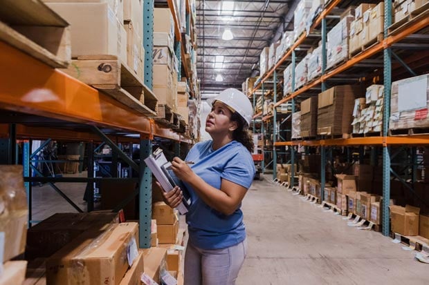 A woman wearing a hard hat and holding a clipboard inspects boxes on shelves in a large warehouse.
