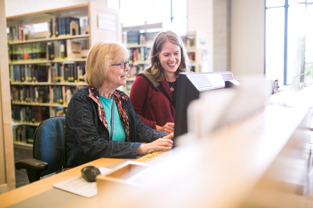Two women are sitting at a computer in a library, with bookshelves in the background. One woman is typing while the other stands beside her, both looking at the screen and smiling.